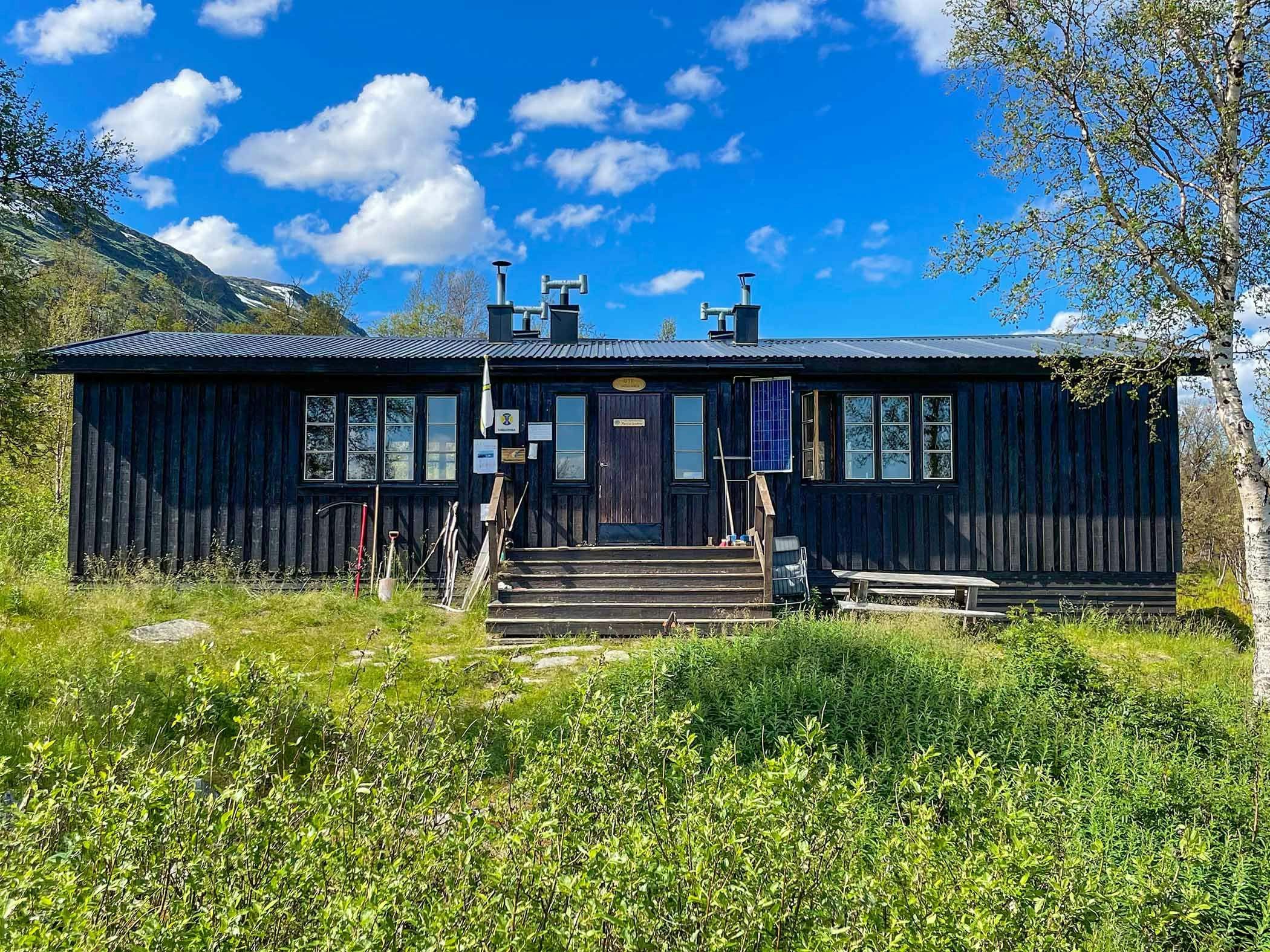 Black mountain cabin in sunshine, surrounded by greenery and blue sky.