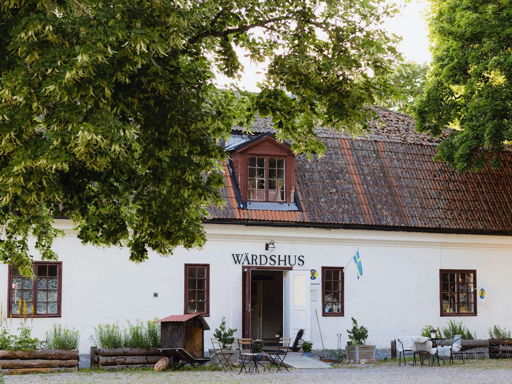 White inn with rustic tiled roof and outdoor seating under large trees.