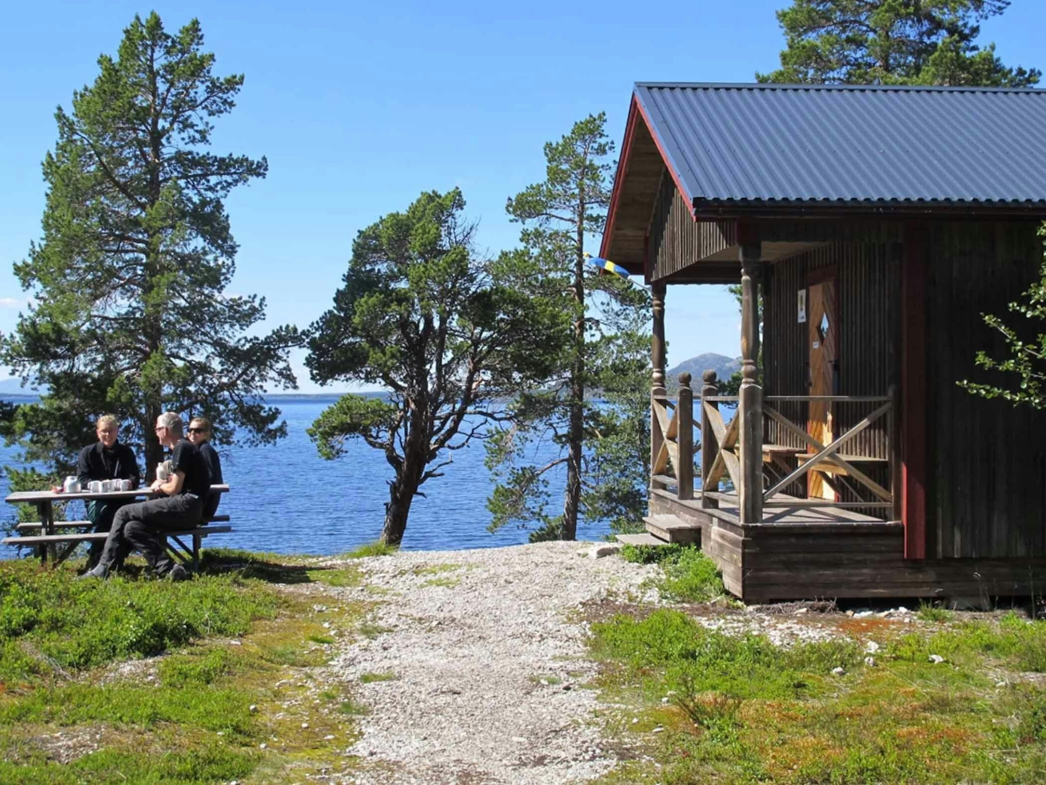 Three people are having coffee at a table next to a log cabin by a blue lake.