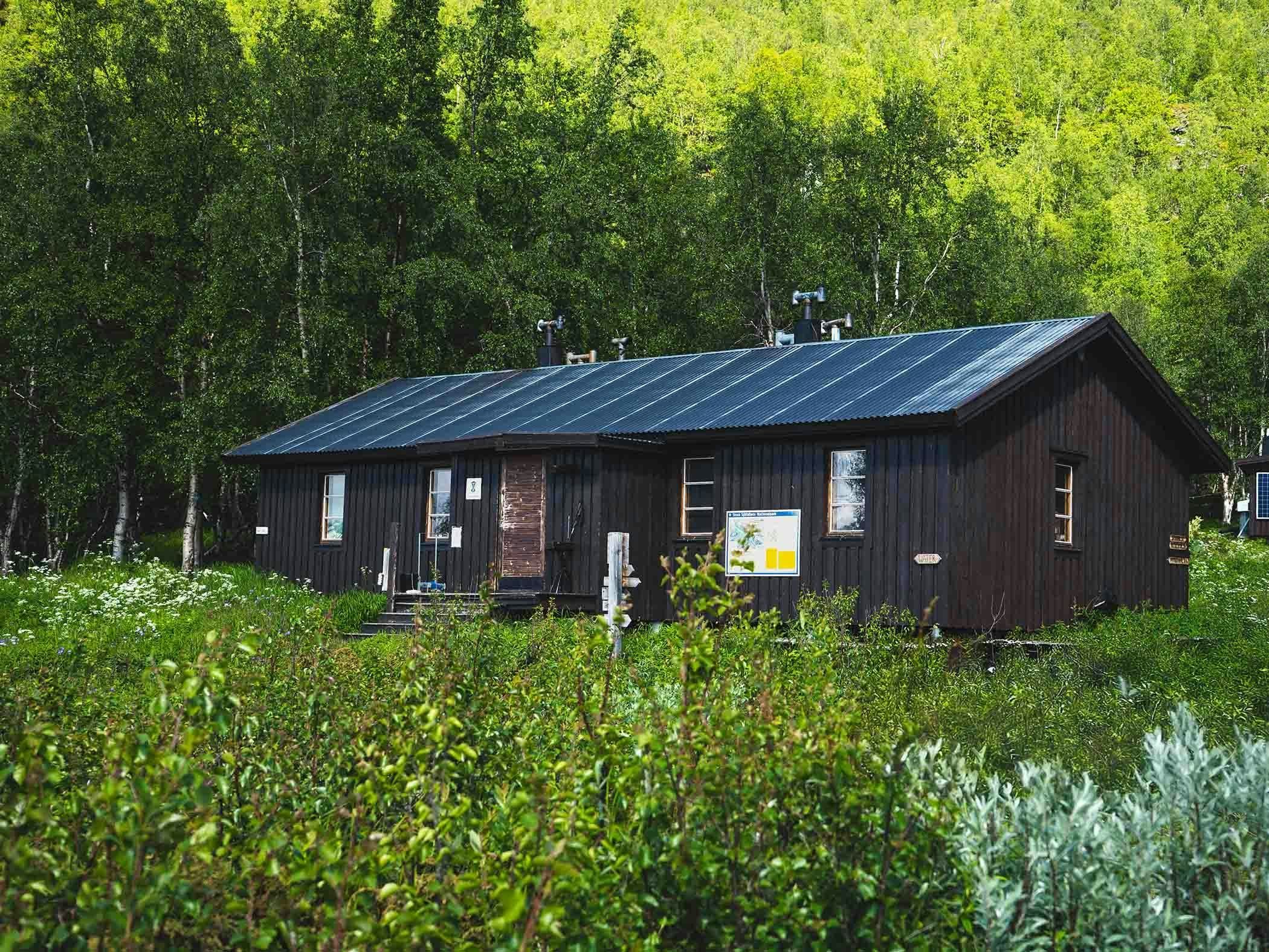 Dark wooden mountain cabin located in a lush valley with a waterfall in the background and signs marking hiking trails.
