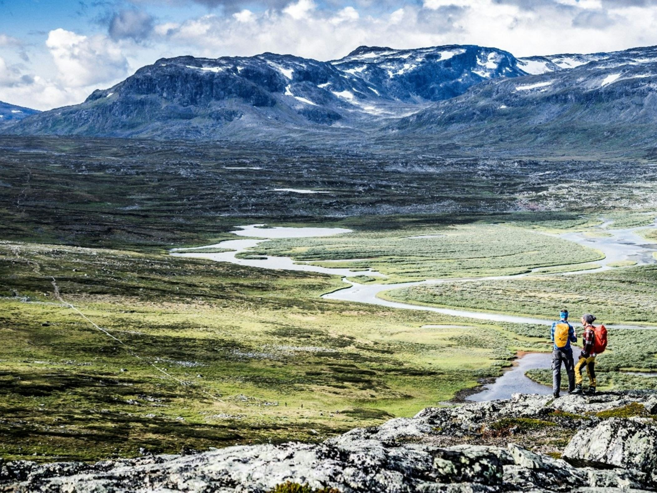 Två vandrare med ryggsäckar blickar ut över ett vidsträckt fjällandskap med slingrande vattendrag och höga berg i bakgrunden.