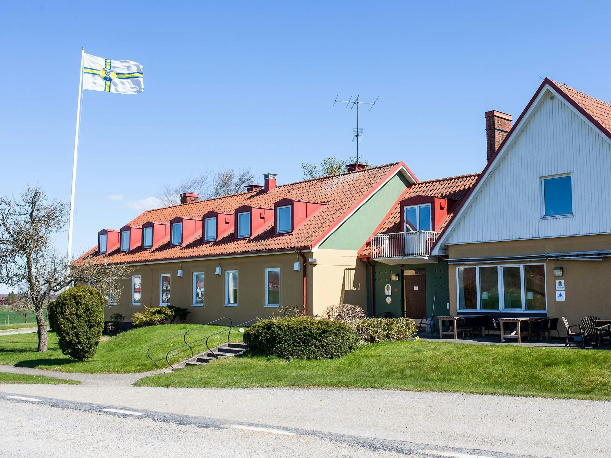 Low yellow building with red dormers and flagpole in a garden setting.