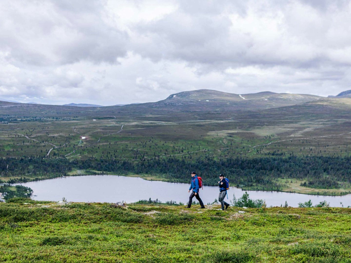 Två personer vandrar. I bakgrunden skymtar sjö och fjäll.