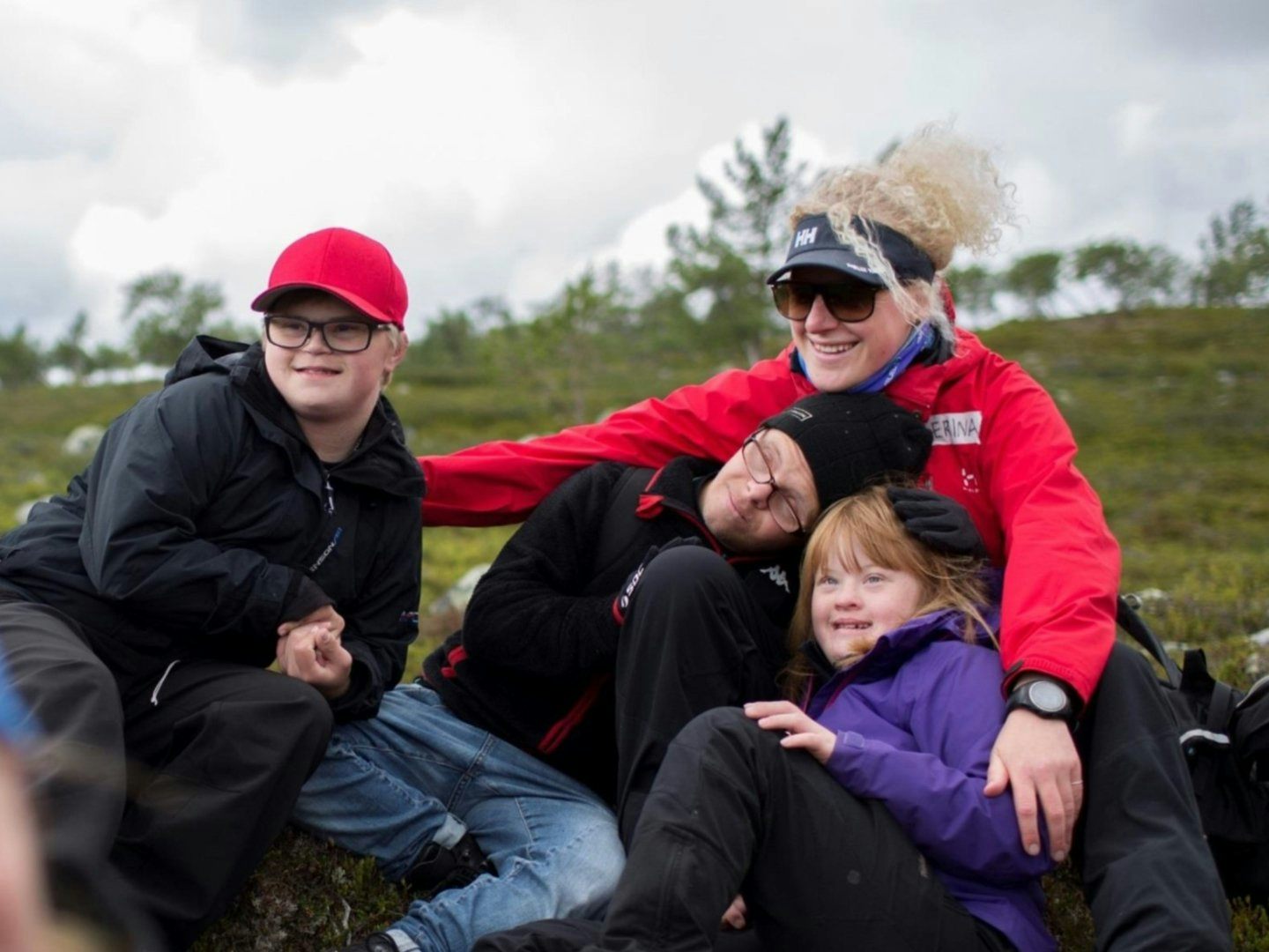En grupp personer sitter tillsammans utomhus på en grönskande plats med träd i bakgrunden, klädda i friluftskläder.