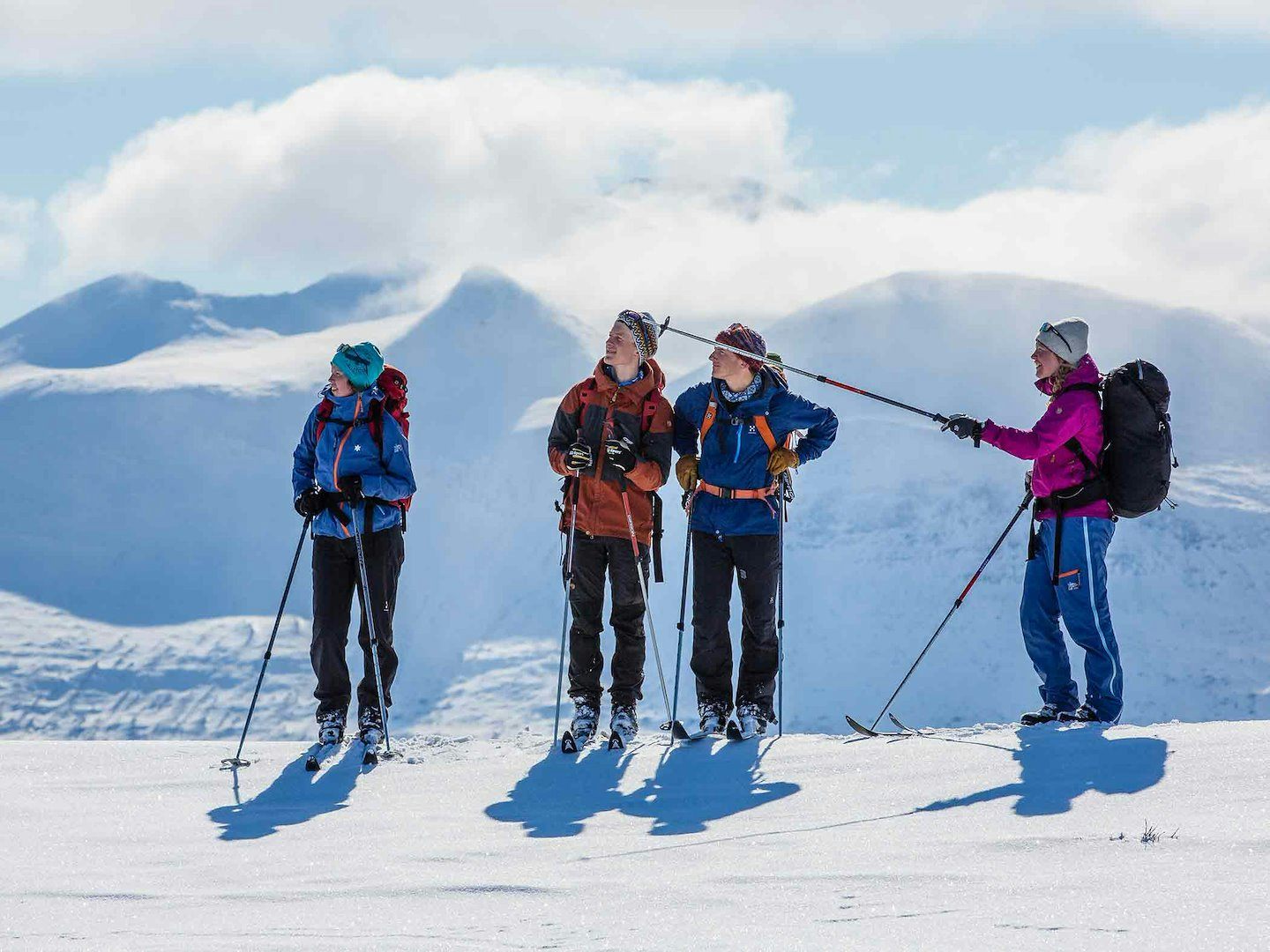 Fyra personer står på fjället med skidor och ryggsäckar, blickar ut över snötäckta berg i bakgrunden.