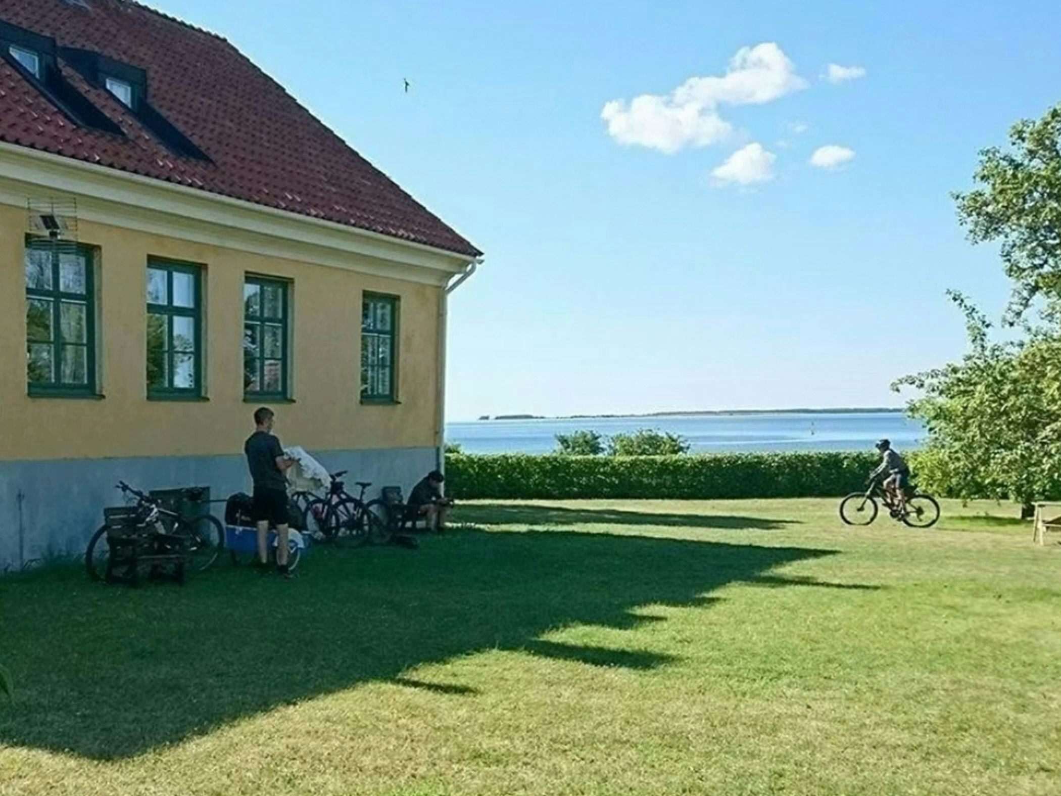 Gulputsad byggnad vid havet, cyklister pausar i trädgården med vy mot öppen horisont.