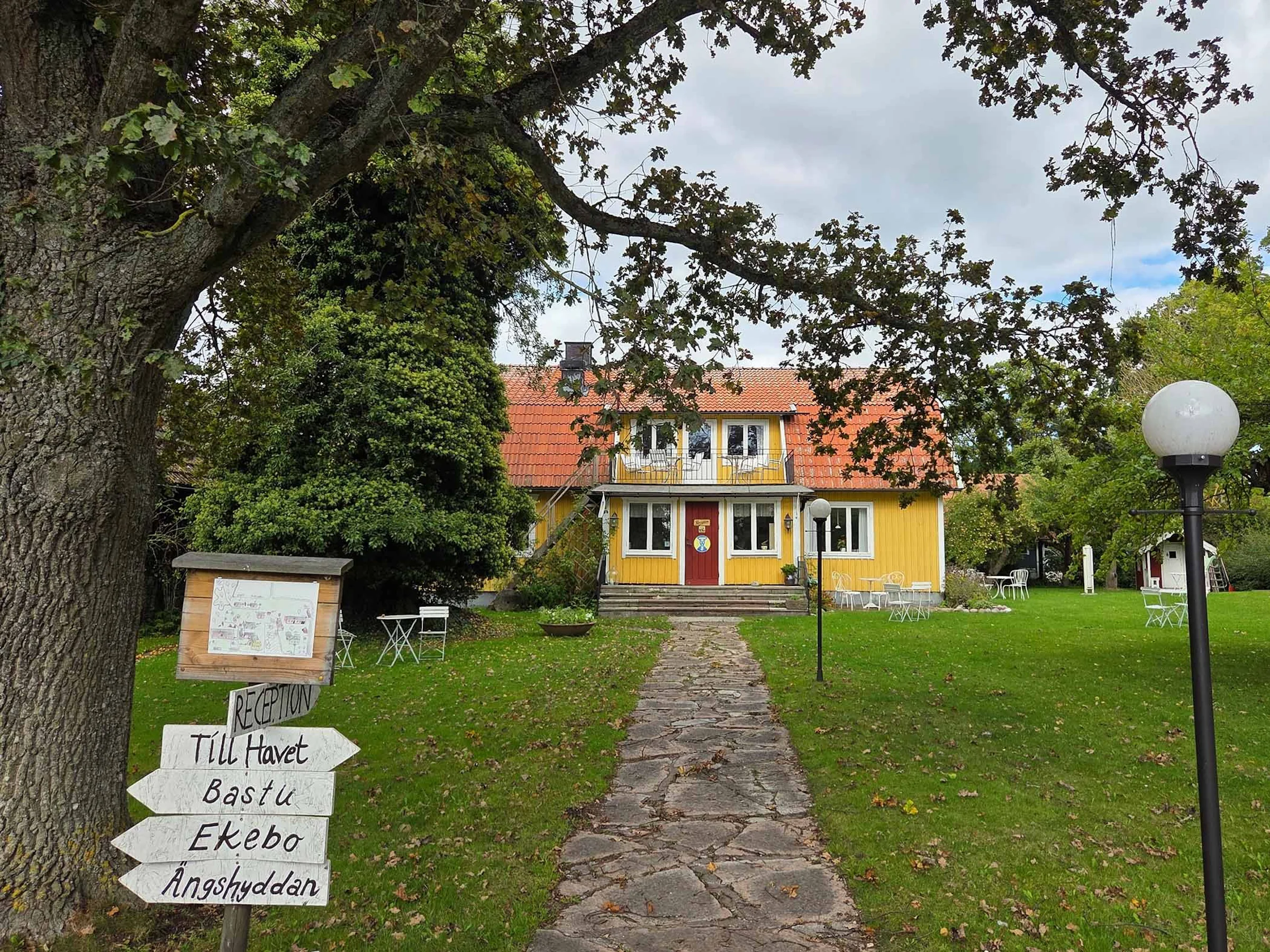 Gul trävilla med rött tegeltak, grön trädgård med trädgårdsmöbler, blå himmel och solsken.