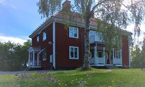 Red wooden villa with white details in a lush garden under a summer sky.