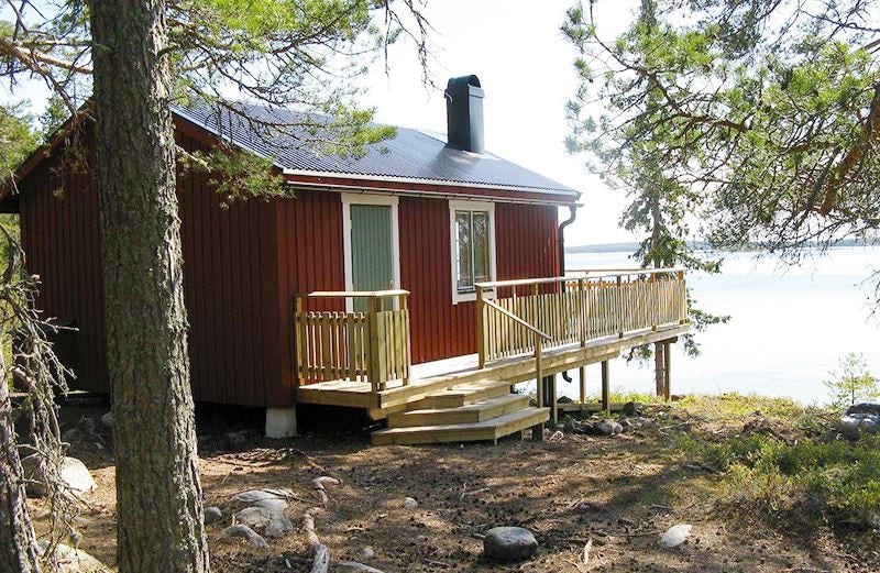 Red cottage with wooden terrace by the water surrounded by pine forest.