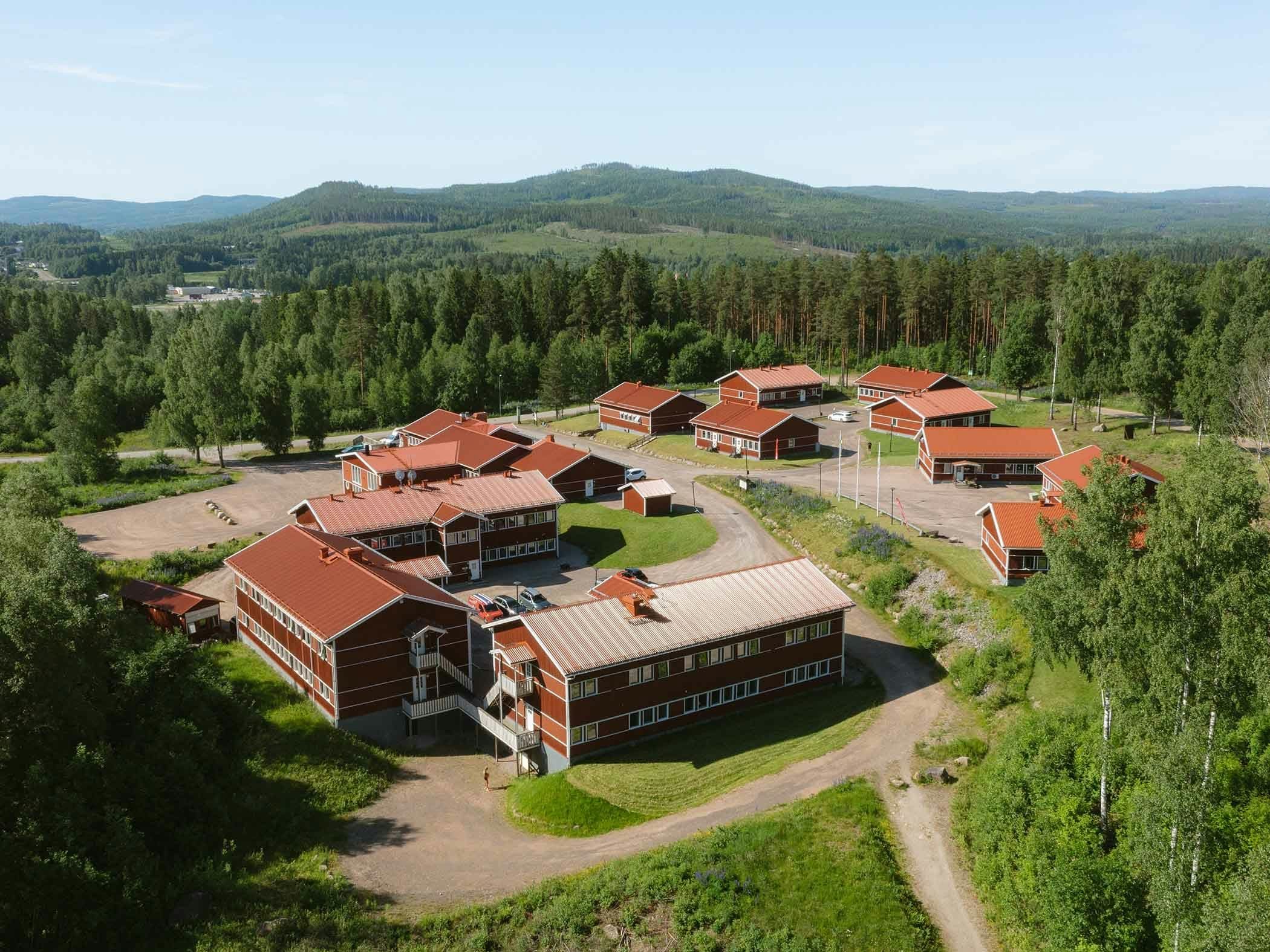 Röd byggnadssamling med röda tak i skogskantad dalgång.